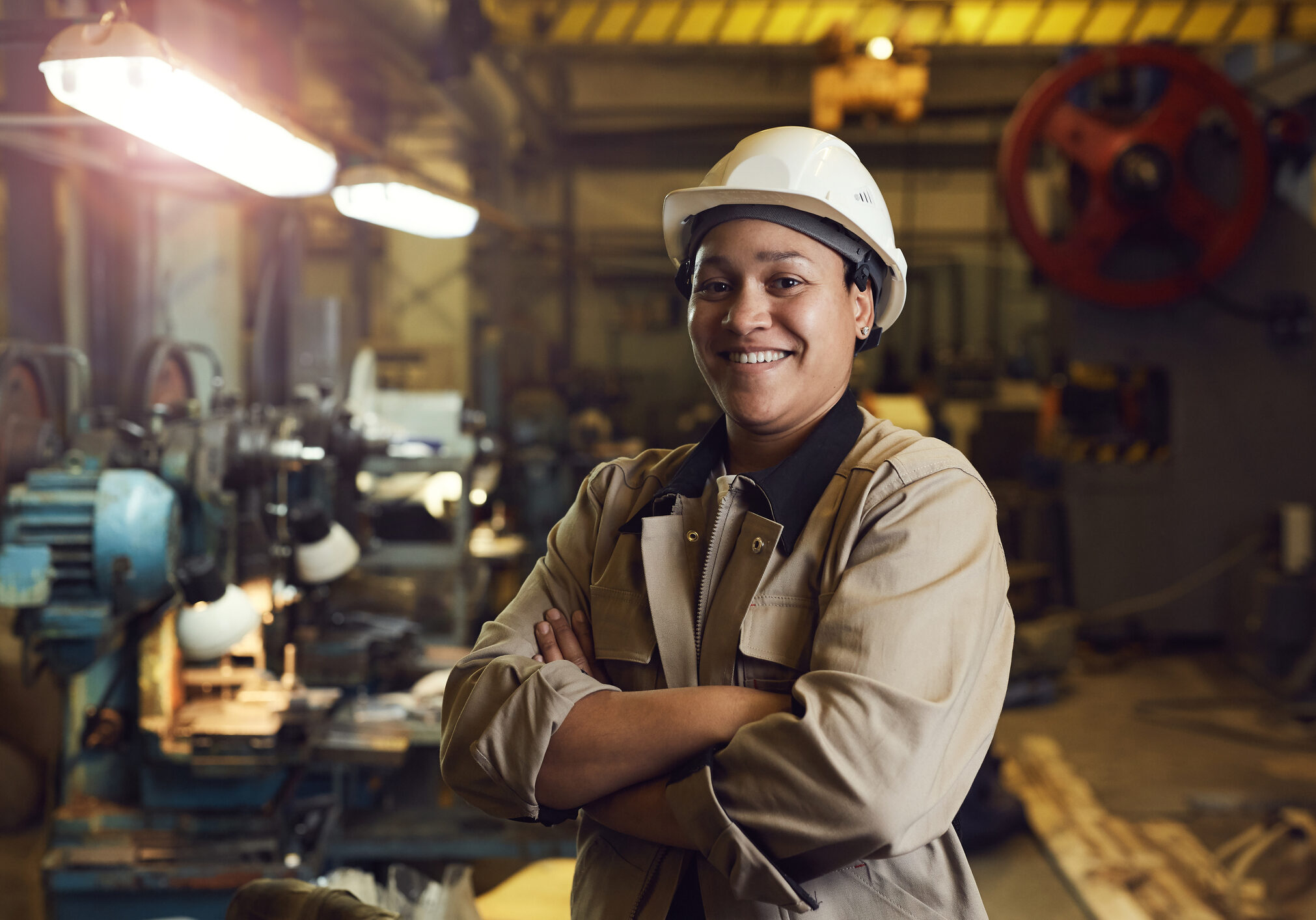 Waist up portrait of mixed-race female worker posing confidently while standing with arms crossed in factory workshop