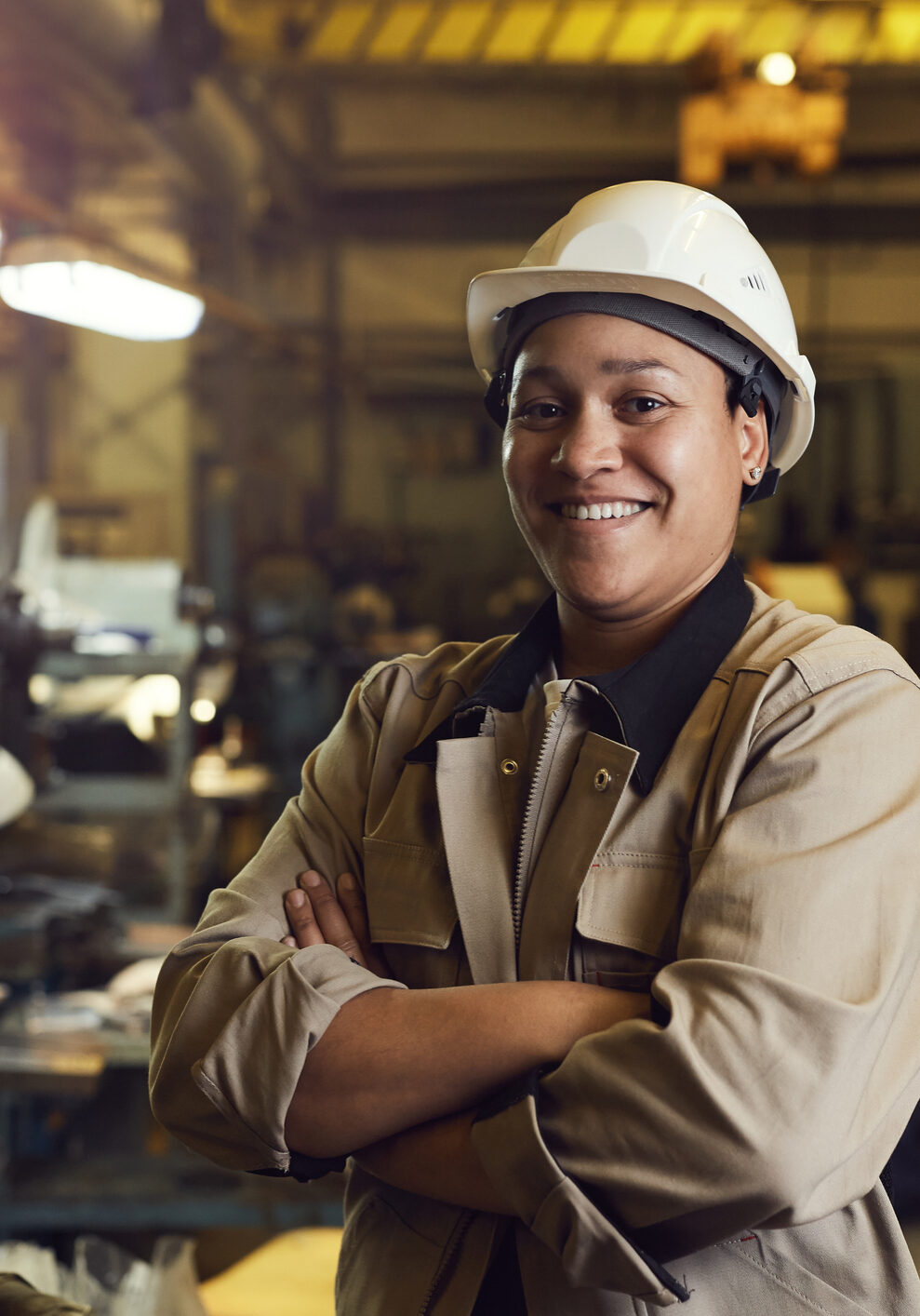 Waist up portrait of mixed-race female worker posing confidently while standing with arms crossed in factory workshop